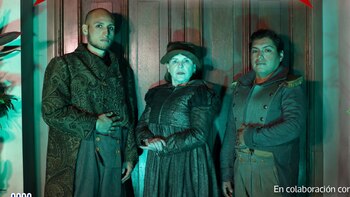 Tres personas, dos hombres y una mujer, visten trajes históricos con un fondo de puerta de madera y plantas