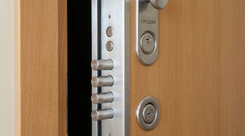 Primer plano de una puerta de seguridad de madera clara abierta, mostrando la barra de metal plateada con cuatro pasadores cilíndricos y un ojo de cerradura.