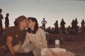 Primer plano de un hombre con gorra besando a una mujer, con el fondo lleno de siluetas de personas y un cielo pálido al anochecer.