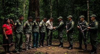 Cinco guardabosques y una mujer indígena son detenidos por cuatro oficiales armados en uniforme, parados en un denso bosque tropical.