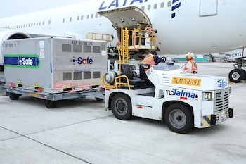 Avión blanco de LATAM en pista, con puerta de carga abierta. Un vehículo remolca un contenedor metálico "Safe". Dos operarios trabajan en la carga