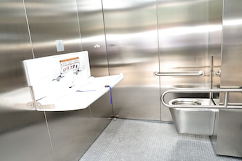 Interior de un baño público moderno con paredes de acero inoxidable, un cambiador de bebé blanco desplegado a la izquierda y un inodoro metálico a la derecha
