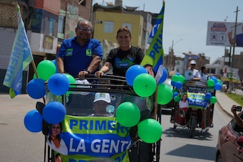 Dos personas sonrientes en un mototaxi decorado con globos verdes y azules y pancartas de "Primero La Gente" en una campaña política por una calle urbana