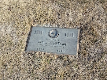 Lápida de Ken Rex McElroy en hierba seca. Muestra su nombre, fechas 1934 y 1981, epitafio, dos libros grabados y un círculo central