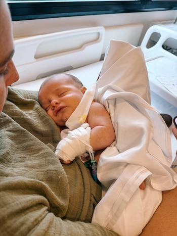 Bebé dormido con vendaje en la muñeca derecha, recostado sobre el hombro de una persona vestida de verde en un entorno hospitalario