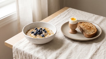 Tazón blanco de avena con arándanos junto a un plato con huevo pasado por agua y tostada sobre un mantel claro.