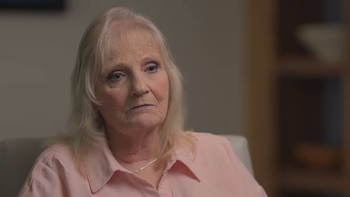 Primer plano de una mujer de edad avanzada con cabello claro y camisa de vestir rosada sobre un fondo desenfocado