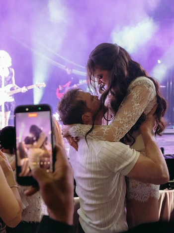 Hombre levanta a mujer en un abrazo, al fondo luces de escenario y músicos. Mano sostiene teléfono móvil grabando a la pareja