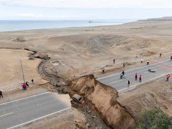 Vista aérea de una carretera asfaltada con un tramo colapsado, un vehículo en el fondo de un barranco, varias personas, un paisaje árido y el mar con un muelle