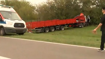 Una ambulancia blanca con rayas rojas y azules y un camión articulado rojo volcado están en la cuneta de una carretera, con un hombre de pie en primer plano