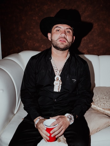 Un hombre con sombrero negro, camisa oscura y un collar con dije religioso está sentado en un sofá blanco sosteniendo un vaso rojo