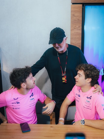Tres hombres, dos sentados con remeras rosadas con logos y uno de pie con gorra negra y remera oscura, en una mesa de madera. Hay un celular en la mesa