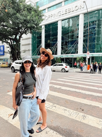 Dos mujeres con gafas de sol sonríen en un paso de peatones frente a un edificio de vidrio con el cartel Shopping Leblon