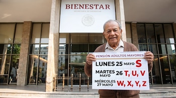 Hombre adulto mayor sonriente sostiene un cartel con el calendario de pagos de Pensión Adultos Mayores frente al edificio de la Secretaría de Bienestar.