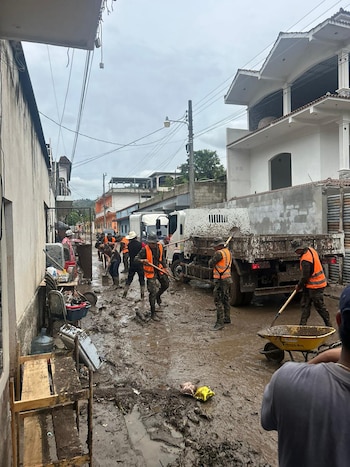 Vista de una calle de tierra cubierta de lodo espeso. Varios hombres con chalecos naranjas y palas limpian la vía junto a un camión blanco con barro y una carretilla