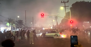 Imagen nocturna de una intersección con un coche gris y negro haciendo derrapes, rodeado de una gran multitud, bajo semáforos en rojo y mucho humo