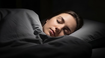 Una mujer con cabello castaño duerme boca arriba sobre una almohada gris, cubierta con una manta gris oscuro en una habitación a oscuras.