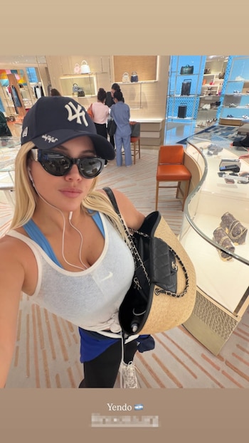Wanda Nara, rubia, con gorra azul de los New York Yankees, gafas de sol negras, camiseta blanca con logo Nike, auriculares, bolso de paja y cartera negra. Fondo de tienda