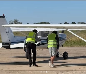 Dos personas con chalecos de seguridad amarillos caminan hacia un pequeño avión blanco y azul en una pista de aterrizaje bajo un cielo despejado