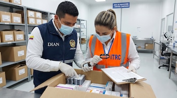 Hombre y mujer con mascarillas y guantes inspeccionan una caja de medicamentos. El hombre usa chaleco DIAN. La mujer, con chaleco ITRC, sostiene lupa y portapapeles.