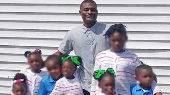 Un hombre negro sonriente con una camisa gris posa de pie junto a un grupo de siete niños con rostros difuminados, delante de una pared blanca de tablillas.