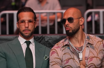 Dois homens sentados; o outro usa óculos, barba, careca, camisa estampada e colar. O outro tem barba, óculos, terno verde, camisa branca e gravata verde