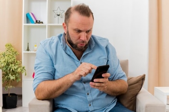 Un hombre barbudo con camisa azul, sentado en un sillón, mira su teléfono móvil con atención, tocando la pantalla con un dedo, con estanterías blancas al fondo