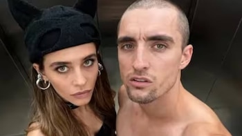 Primer plano de una mujer con gorro negro de orejas y aretes de aro y un hombre sin camisa con pelo rapado. Fondo oscuro