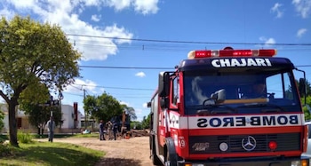 Primer plano de un camión de bomberos de Chajarí con luces rojas intermitentes estacionado en una calle soleada, con árboles y personas al fondo
