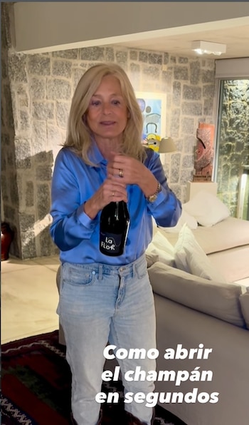 Cecilia Zuberbühler, mujer rubia de mediana edad, de pie en un salón, viste camisa azul y jeans, sosteniendo y abriendo una botella de espumante negro