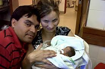 Primer plano de un hombre y una mujer sosteniendo a un bebé recién nacido envuelto en una manta blanca; la mujer sonríe a la cámara