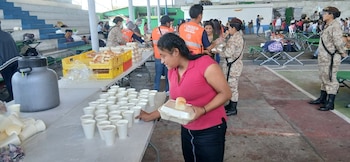Múltiples personas en un albergue; mesas con vasos de bebida, panes y contenedores de comida; personal con chalecos COMRED y militares asisten
