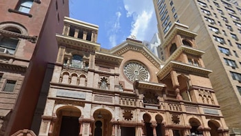 Vista frontal de un edificio antiguo de ladrillo con arcos ornamentados, un rosetón y texto en hebreo, flanqueado por rascacielos bajo un cielo azul