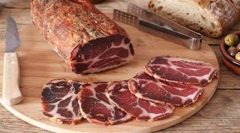 Primer plano de un embutido curado rojizo y veteado, parcialmente rebanado, sobre una tabla de madera con un cuchillo y tenazas. Al fondo, pan y aceitunas.