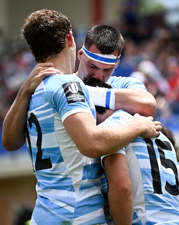 Tres jugadores de rugby se abrazan en el campo de juego. Llevan camisetas con franjas celestes y blancas y los números 12 y 15. Uno usa una vincha azul y blanca