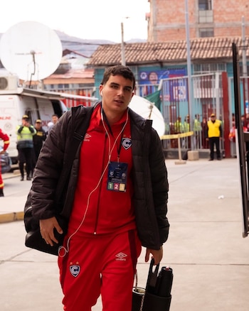 Hombre joven con chaqueta negra y chándal rojo del equipo Cienciano, lleva auriculares, una credencial, una bolsa y un termo, con vehículos de televisión al fondo