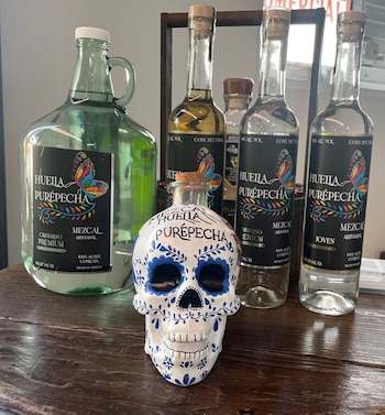 Seis botellas de vidrio, incluyendo una en forma de calavera, con etiquetas de mezcal Huella Purépecha se disponen sobre una mesa de madera