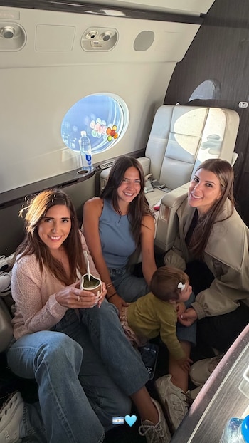 Tres mujeres y un bebé sonríen dentro de un avión. Una mujer sostiene un mate. Se observan asientos de cuero y una ventana con botellas de agua