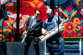 Dos hombres en un escenario tocando instrumentos; uno con guitarra eléctrica azul, otro con keytar. Fondo con pantalla LED de patrones coloridos