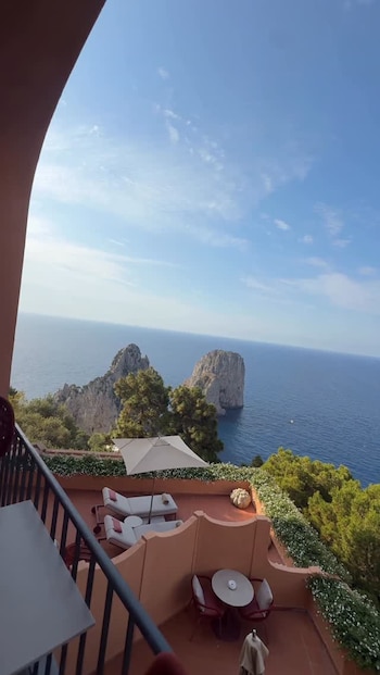 Vista aérea desde un balcón con una terraza amueblada, mirando hacia el mar azul claro y tres grandes formaciones rocosas cubiertas de vegetación en la distancia