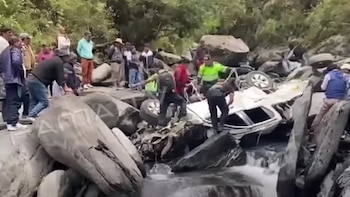 Una camioneta blanca volcada y destruida entre rocas y agua, rodeada por rescatistas y personas que observan