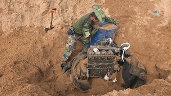 Um homem mascarado em uma caixa de areia ao lado de um motor sujo e um barril azul, com uma pá cravada na areia ao fundo.