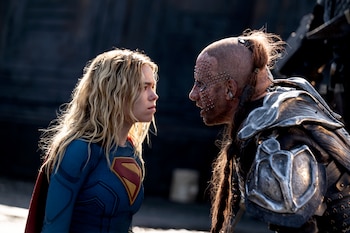 Milly Alcock como Supergirl con traje azul y capa roja frente a Matthias Schoenaerts con armadura metálica, cabeza rapada y piercings faciales