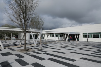 Edificio moderno blanco con ventanas amplias y un patio exterior pavimentado con patrones geométricos en blanco y negro; dos personas y un perro caminan cerca, bajo un cielo nublado