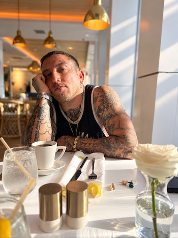 Un hombre con múltiples tatuajes apoya la cabeza en la mano frente a una mesa con una taza de café, cubiertos, agua y varias cápsulas