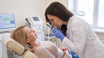 Una médica con bata blanca y guantes azules utiliza un dermatoscopio para observar el rostro de una mujer recostada en una camilla.