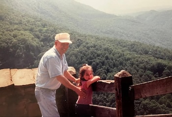 Un hombre adulto con gorra beige y camiseta clara junto a una niña con camiseta rosa apoyada en una baranda de madera. Al fondo, montañas cubiertas de árboles
