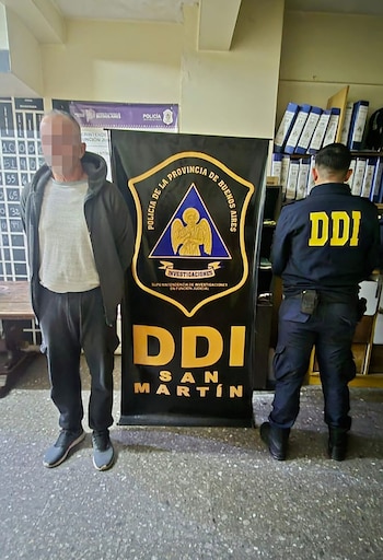 Un hombre con rostro pixelado, vestido con ropa oscura, de pie junto a un cartel de la DDI San Martín, y un oficial de espaldas