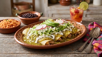 Enchiladas de pollo en salsa verde con lechuga, crema, queso Cotija, rábano y aguacate en plato artesanal, acompañadas de arroz, frijoles y agua fresca.