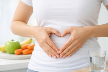 Manos sobre abdomen con camiseta blanca formando un corazón. Al fondo, desenfocado, se ven manzana verde, plátanos, zanahorias y un vaso de agua.
